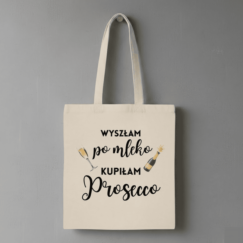 Beżowa materiałowa torba z czarnym napisem: ‘Wyszłam po mleko, kupiłam Prosecco’, ozdobiona rysunkami kieliszka z winem i butelki Prosecco